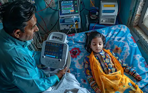 A concerned man monitors a young girl lying in a hospital bed, connected to medical equipment, highlighting the urgent need for care and the hope your donation can bring.