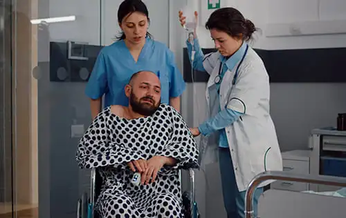 A man sitting in a wheelchair being cared for by a female doctor holding a saline bag and a female nurse offering support, symbolizing compassion and hope through medical care.
